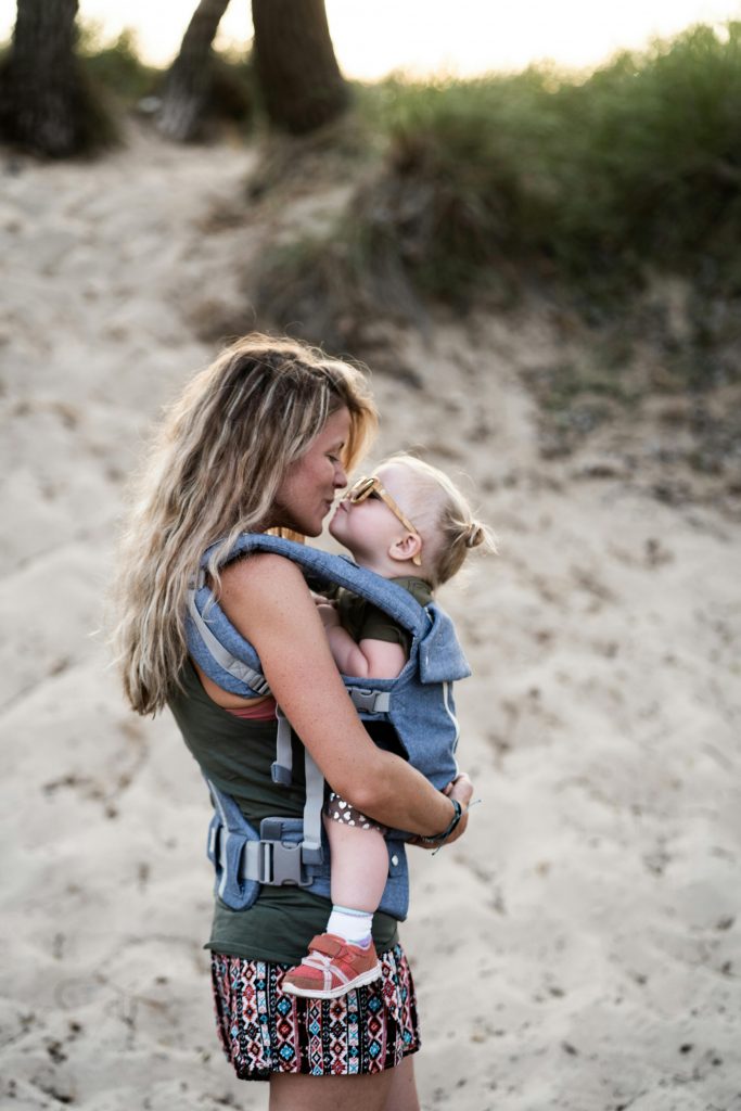 A loving mother kisses her toddler in a baby carrier on a sandy beach, capturing a tender parenting moment.